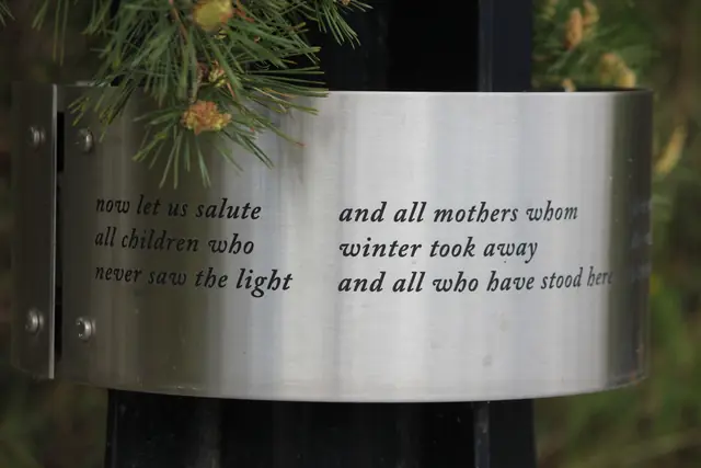 An engraved metal plaque with a poem is mounted on a post beneath pine branches.
