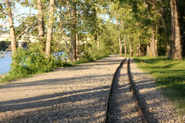 A rusted rail curves along a gravel path beside a river with a bridge in the distance.