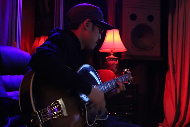A man in a cap plays an archtop guitar in a dim room lit by a red lamp and blue light.