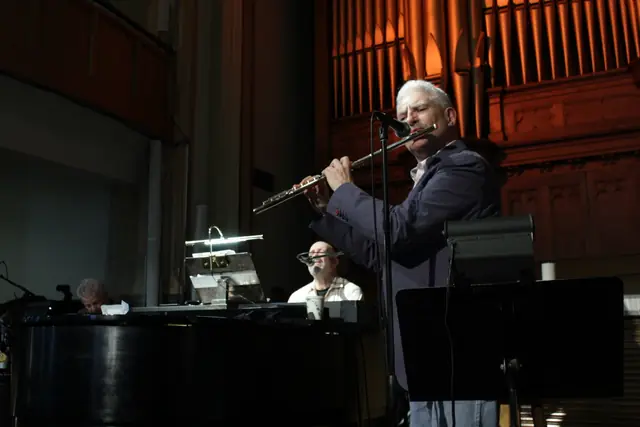 A man plays a flute into a microphone on a stage with organ pipes behind him.