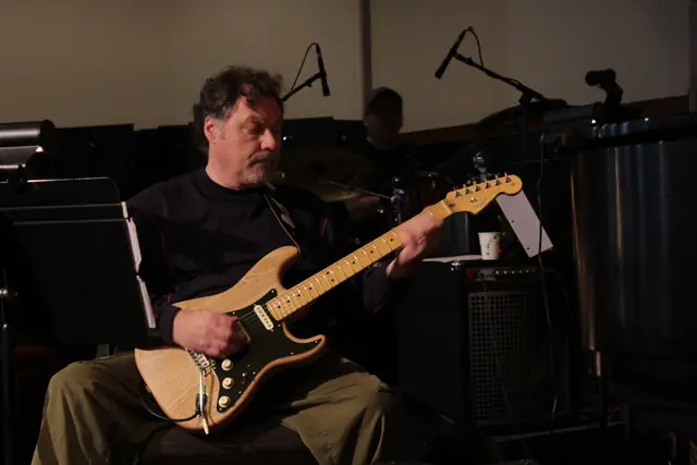 A man plays an electric guitar on a dimly lit stage with a drummer behind him.