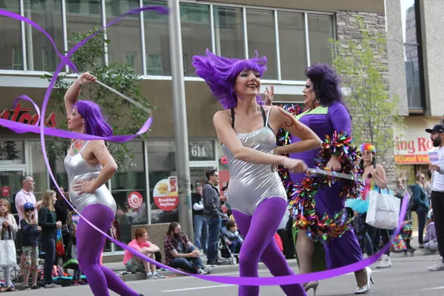 Three dancers in purple wigs and costumes perform in the street with a ribbon swirling overhead.