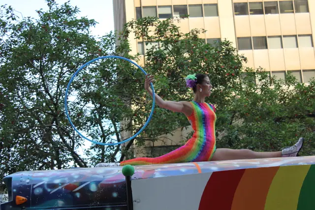 A performer in a rainbow bodysuit poses on a parade float holding a large hoop overhead.