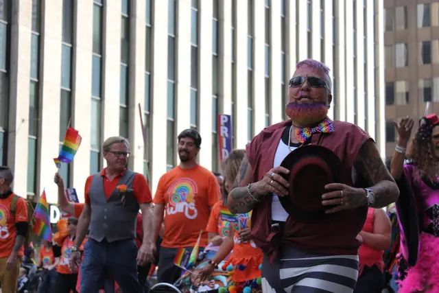 A man with a pink-dusted beard and a rainbow bow tie walks the parade route holding a hat.