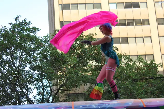 A performer with colourful hair stands on a parade float as a pink scarf billows behind her.