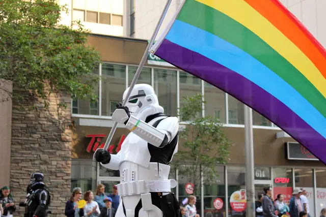 A person in a stormtrooper costume holds up a large rainbow flag on a city street.