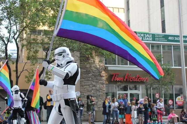 A person in a stormtrooper costume carries a rainbow flag down a city street.