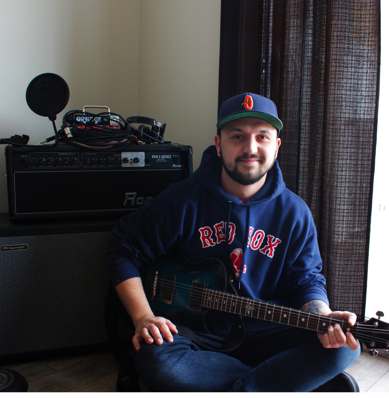 Michael Williams seated with an electric guitar in his home studio, amplifiers and a microphone behind him.