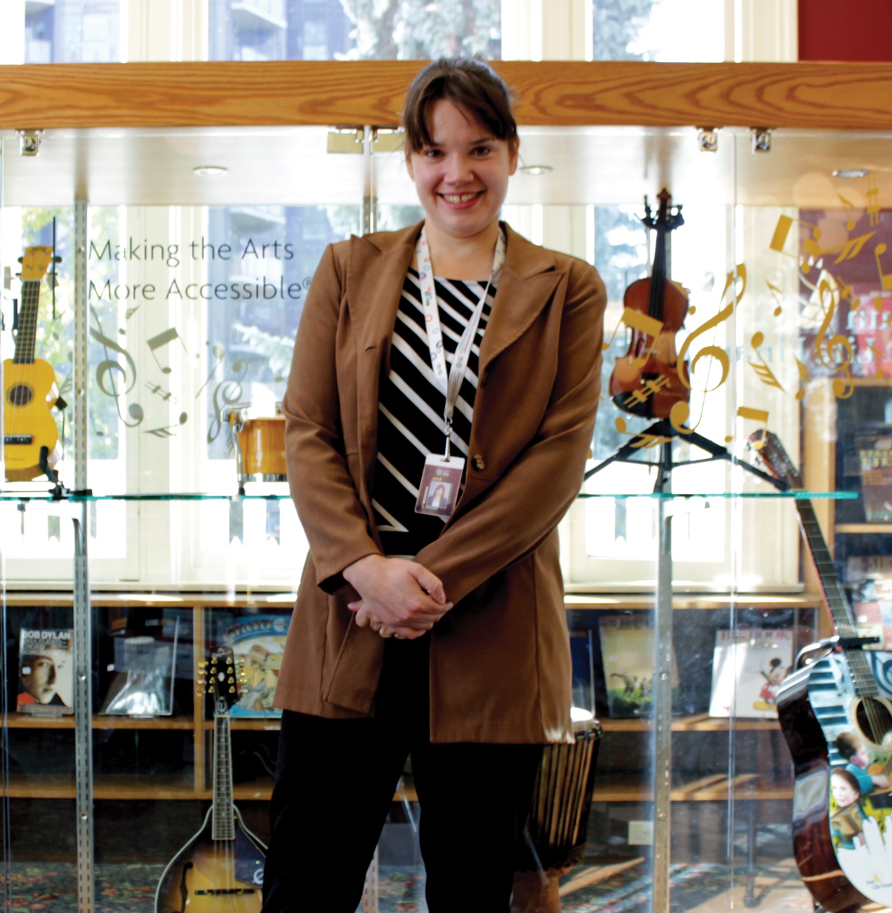 Julia Harrington standing in front of a glass display of instruments at Memorial Park Library, under a sign reading “Making the Arts More Accessible”.