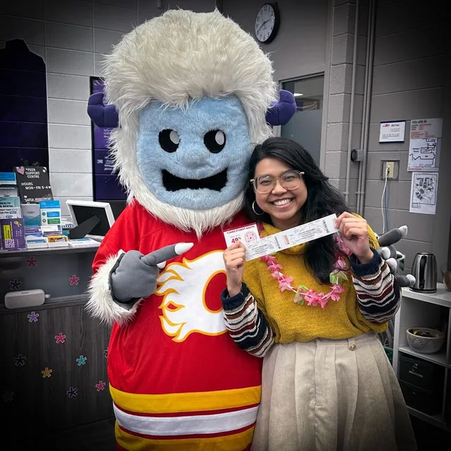 Tedi the Yeti in a Calgary Flames jersey pointing at a smiling student holding a Flames Student Nights ticket